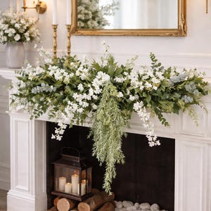 Può includere: Un caminetto bianco adornato con una composizione floreale di fiori bianchi e verde. Uno specchio con cornice dorata è appeso sopra il caminetto. Una lanterna con candele, tronchi e pietre è nel camino.