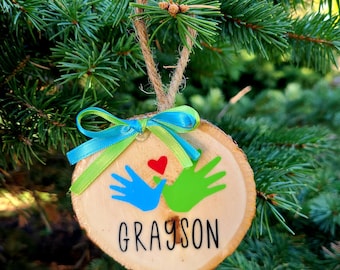 May include: A wooden Christmas ornament with a blue and green handprint design and a red heart. The ornament is decorated with a blue and green ribbon and the name "GRAYSON" is written on the front.
