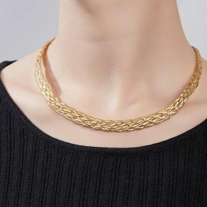 Könnte beinhalten: Eine goldfarbene Kettenkette mit geflochtenem Design. Die Kette wird von einer Person mit einem schwarzen Oberteil getragen.