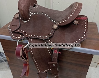 May include: A brown leather saddle with white stitching and silver accents. The saddle is on a wooden surface and has a stirrup on the left side. The saddle is labeled "Equistallion Saddlery Co."