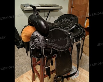 May include: A black leather western saddle with a tooled design and a white stitching detail. The saddle is sitting on a wooden stool.