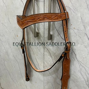 Può includere: Una briglia per cavalli in pelle marrone con intricate lavorazioni floreali. La briglia ha una sottogola nera e una fronte con un dettaglio di cucitura decorativo.