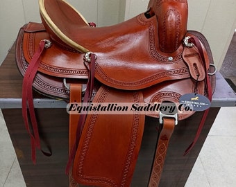 May include: A brown leather saddle with intricate stitching and a tooled design. The saddle has a high cantle and a horn with a leather wrap. The saddle is sitting on a wooden surface.