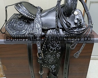May include: A black leather saddle with silver floral tooling and a black leather breast collar. The saddle is on a wooden surface.