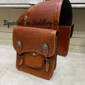 Peut inclure: Une sacoche de selle en cuir marron avec des détails complexes et des accents argentés. Le sac a un rabat avec une boucle et une petite poche sur le devant. La sacoche de selle est étiquetée "Equistallion Saddlery Co."