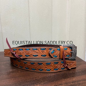 Puede incluir: Un cinturón de cuero marrón con costuras turquesa y una hebilla plateada. El cinturón está decorado con un patrón floral y el texto "EQUISTALLION SADDLERY CO."