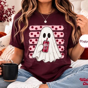 May include: A maroon t-shirt featuring a graphic of a white ghost holding a red soda can with the text Dr Pepper 23. The ghost is positioned in front of a background of five horizontal pink brush strokes decorated with dark pink polka dots.