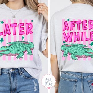 K&ouml;nnte beinhalten: Hellgraues T-Shirt mit rosa und wei&szlig;en Streifen. Auf der Vorderseite steht das Wort &bdquo;LATER&ldquo; in Rosa, ein gr&uuml;nes Alligator und gr&uuml;ne Sterne. Auf der R&uuml;ckseite steht &bdquo;AFTER WHILE&ldquo; und ein gr&uuml;nes Alligator. Ein lustiges, l&auml;ssiges Oberteil.