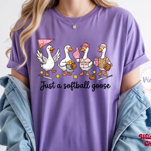 May include: A purple t-shirt featuring a cartoon design of four geese playing softball. The geese are wearing softball gear and holding equipment. The text "Just a softball goose" is printed below the geese.