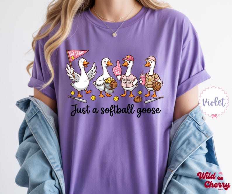 May include: A purple t-shirt featuring a cartoon design of four geese playing softball. The geese are wearing softball gear and holding equipment. The text "Just a softball goose" is printed below the geese.