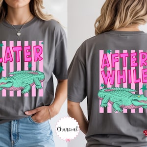 K&ouml;nnte beinhalten: Anthrazitgraues T-Shirt mit einem rosa-wei&szlig; gestreiften Rechteck-Design. Auf der Vorderseite des Shirts steht "LATER" &uuml;ber einer gr&uuml;nen Alligator-Illustration. Auf der R&uuml;ckseite steht "AFTER WHILE" &uuml;ber einer gr&uuml;nen Alligator-Illustration.