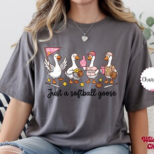 May include: Charcoal gray t-shirt featuring a cartoon illustration of four geese dressed in softball gear. The geese are holding a pennant, a foam finger, a softball, and a drink. The text "Just a softball goose" is below the geese.