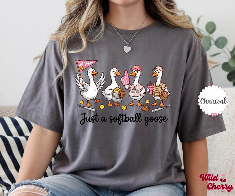 May include: Charcoal gray t-shirt featuring a cartoon illustration of four geese dressed in softball gear. The geese are holding a pennant, a foam finger, a softball, and a drink. The text "Just a softball goose" is below the geese.
