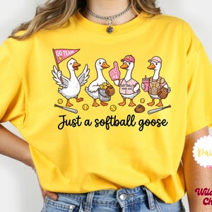 May include: Yellow t-shirt featuring a whimsical illustration of four geese dressed for softball. The geese are holding a pennant, a bucket of balls, a foam finger, and a drink. Text on the shirt reads "Just a softball goose."