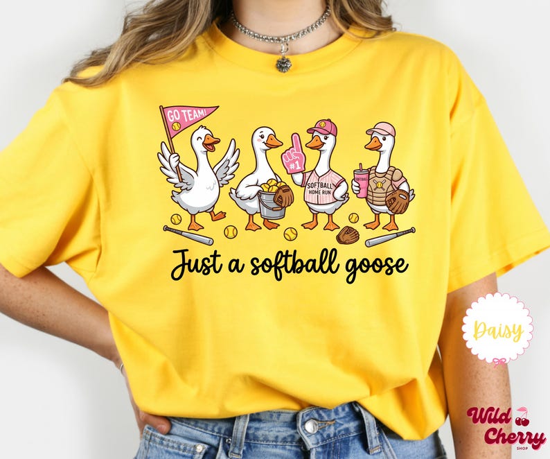 May include: Yellow t-shirt featuring a whimsical illustration of four geese dressed for softball. The geese are holding a pennant, a bucket of balls, a foam finger, and a drink. Text on the shirt reads "Just a softball goose."
