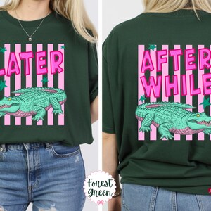 K&ouml;nnte beinhalten: Forstgr&uuml;nes T-Shirt mit einem rosa und wei&szlig; gestreiften Rechteck-Design. Auf der Vorderseite des Shirts steht "LATER" in rosa Buchstaben &uuml;ber einem t&uuml;rkisfarbenen Alligator. Auf der R&uuml;ckseite steht "AFTER WHILE" &uuml;ber einem t&uuml;rkisfarbenen Alligator.