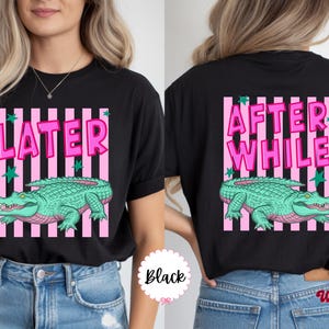 K&ouml;nnte beinhalten: Schwarzes T-Shirt mit einem Grafikdesign, das ein gr&uuml;nes Alligator zeigt und den Text "LATER" auf der Vorderseite und "AFTER WHILE" auf der R&uuml;ckseite. Das Design umfasst rosa Streifen und gr&uuml;ne Sterne.