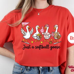 May include: Red t-shirt with a graphic of four cartoon geese playing softball. The geese are wearing baseball caps, holding a pennant, a foam finger, a drink, and a catcher's mitt. The text "Just a softball goose" is below the geese.