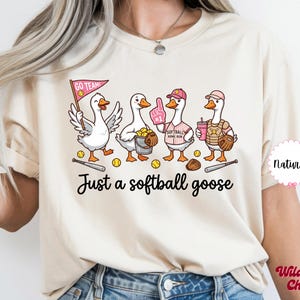 May include: Cream-colored t-shirt featuring a whimsical illustration of four geese dressed as softball players. The geese are holding a bat, a glove, and a drink. The text "Just a softball goose" is printed below the illustration.