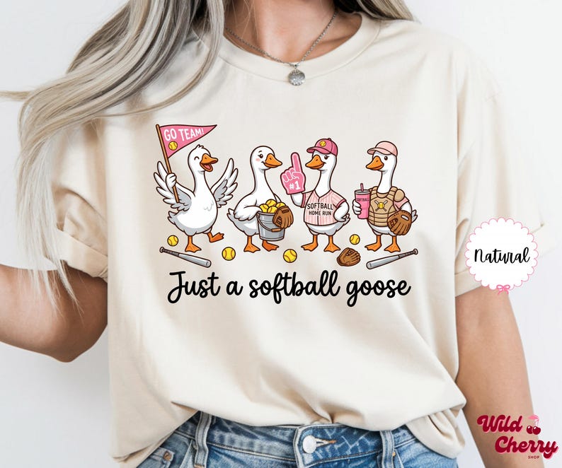 May include: Cream-colored t-shirt featuring a whimsical illustration of four geese dressed as softball players. The geese are holding a bat, a glove, and a drink. The text "Just a softball goose" is printed below the illustration.