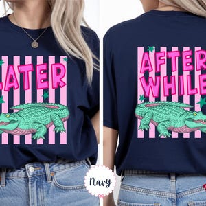K&ouml;nnte beinhalten: Marineblaues T-Shirt mit einer Grafik eines gr&uuml;nen Alligators und dem Text "LATER" auf der Vorderseite. Auf der R&uuml;ckseite steht "AFTER WHILE" und die gleiche Alligatorengrafik. Das Design umfasst rosa Streifen und gr&uuml;ne Sterne.