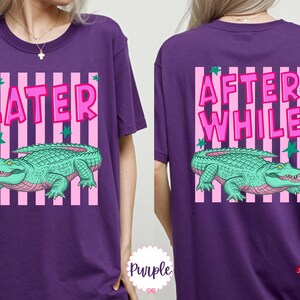 K&ouml;nnte beinhalten: Lila T-Shirt mit einem vorderen Aufdruck &bdquo;LATER&ldquo; und einem hinteren Aufdruck &bdquo;AFTER WHILE&ldquo;. Beide Grafiken zeigen ein gr&uuml;nes Alligator &uuml;ber rosa und wei&szlig;en Streifen, mit gr&uuml;nen Sternen.