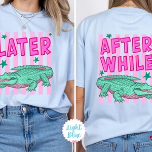 K&ouml;nnte beinhalten: Hellblaues T-Shirt mit rosa und wei&szlig;en Streifen. Auf der Vorderseite steht das Wort "LATER" in Pink, ein gr&uuml;nes Alligator und gr&uuml;ne Sterne. Auf der R&uuml;ckseite stehen die Worte "AFTER WHILE" und ein gr&uuml;nes Alligator.