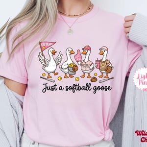 May include: Light pink t-shirt featuring a cartoon illustration of four geese dressed in softball gear. The geese are holding a flag, a bucket of balls, a foam finger, a drink, and a glove. The text "Just a softball goose" is below the illustration.