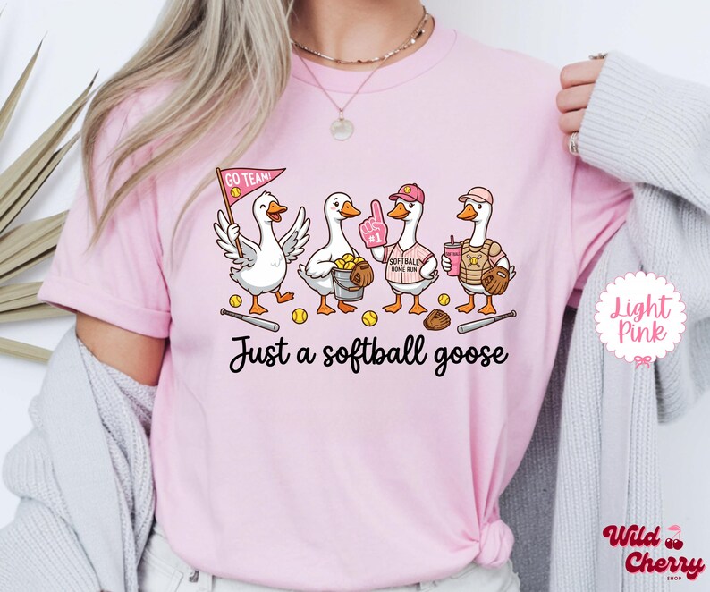 May include: Light pink t-shirt featuring a cartoon illustration of four geese dressed in softball gear. The geese are holding a flag, a bucket of balls, a foam finger, a drink, and a glove. The text "Just a softball goose" is below the illustration.