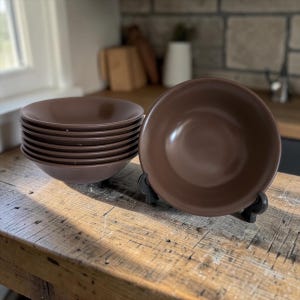 May include: A stack of seven round, solid brown ceramic bowls with a smooth, matte finish. One bowl is positioned upright on a black display stand in front of the stack, showcasing the deep, circular interior and the consistent dark brown colour.