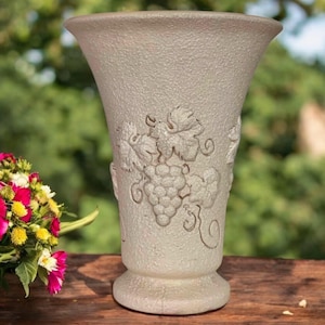 May include: A frosted glass vase with a textured surface and a flared top. The vase features an embossed grape and vine design. The vase is sitting on a wooden surface with a blurred green background.