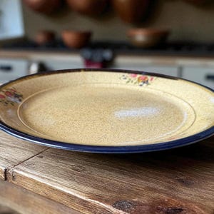 May include: A round, ceramic serving plate with a speckled tan centre and a dark blue rim. The plate has a floral design in the upper left corner. The plate is sitting on a wooden table.