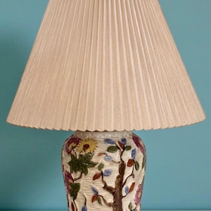 May include: A table lamp with a beige pleated lampshade. The ceramic base is decorated with a hand-painted floral and tree design in multiple colours. The lamp sits on a light blue surface against a matching background.