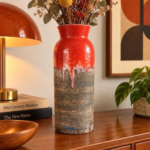 May include: A tall cylindrical ceramic vase featuring a vibrant red glazed upper section that drips down onto a textured, dark grey, and earthy brown base. The vase is filled with a dried floral arrangement consisting of yellow billy balls and greenery.