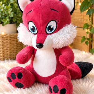 May include: A plush fox toy featuring vibrant red fur, a white chest patch, and white fluffy cheek tufts. The fox has large blue eyes with black eyelashes, a black nose, and black-tipped ears. Its paws have black pads, and it has a black tail.