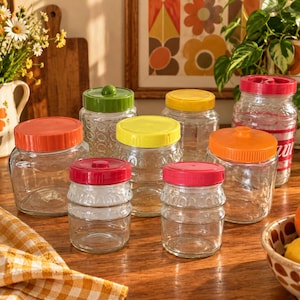 May include: A collection of clear glass storage jars with colourful plastic lids in shades of orange, green, yellow, and red. The jars feature various textured patterns on the glass surfaces, including circular and ribbed designs, arranged on a wooden surface.