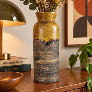 May include: A tall cylindrical ceramic vase featuring a two-tone glaze. The upper portion is a smooth, mustard-yellow finish, while the lower section displays a dark, textured, charcoal-grey surface with horizontal ridges and subtle earthy speckles.