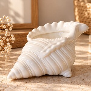 May include: A white ceramic seashell-shaped planter or decorative bowl featuring a glossy finish, intricate ribbed textures, and a scalloped edge. The object is designed with a tapered spiral end and small feet for stability on a flat surface.