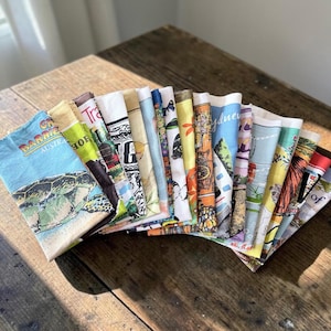 May include: A collection of folded rectangular fabric tea towels arranged in a fan shape. Each towel features distinct colourful graphic prints, including illustrations of a sea turtle, Australian landmarks, floral patterns, and various travel-themed motifs.