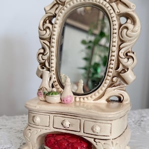 May include: A cream-coloured miniature vanity set with an oval mirror, ornate frame, and three small drawers. It features a red cushioned stool and small decorative bottles. This vintage-style vanity is a charming decorative piece.