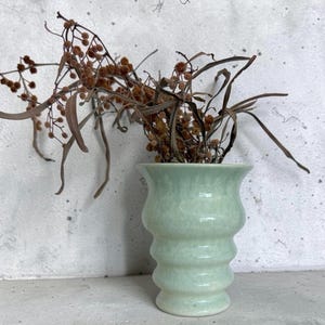 May include: A light mint green ceramic vase with a tiered, ribbed silhouette and a flared rim. The vase contains a cluster of dried brown botanical stems with small round seed pods and long, thin, drooping leaves extending outwards.