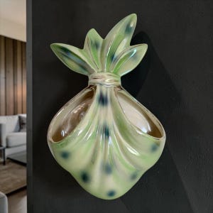 May include: A wall-mounted ceramic planter shaped like a tied fabric bundle. The piece features a glossy finish in shades of pale green with darker green and blue accents, including a gathered top section and a wide, open pocket for plants.