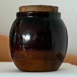 May include: A small, round, brown ceramic jar with a cork stopper. The jar has a glossy finish, with a darker brown colour at the top that fades to a lighter brown at the base. The cork stopper is a light brown colour.