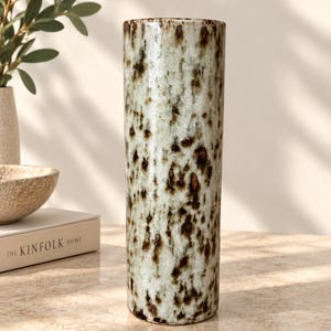 May include: A tall, cylindrical ceramic vase featuring a mottled glaze with a cream-coloured base and dark brown, speckled markings throughout the surface. The vase has a smooth, straight-sided profile with a flat top opening and a flat base.
