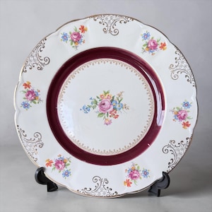 May include: A white porcelain plate with a scalloped edge and a claret-coloured centre ring. The plate features floral bouquets in pink, blue, and yellow, and gold filigree details. The plate is displayed on a black stand.
