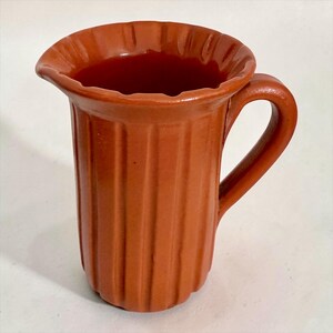 May include: A terracotta-coloured ceramic pitcher featuring a vertical ribbed pattern along the body, a curved handle on the side, and a wide, scalloped rim with a small pouring spout. The surface has a smooth, uniform finish throughout the piece.
