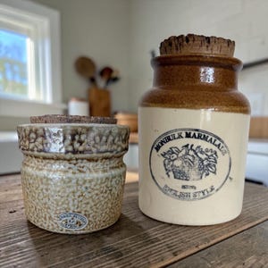May include: A short, textured beige ceramic jar with a cork lid and a small oval stamp, next to a taller, smooth cream-coloured ceramic jar with a brown rim, a cork lid, and a black circular label reading Monbulk Marmalade English Style.