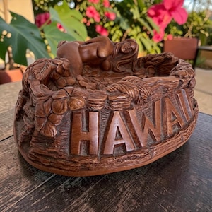 May include: A brown carved wooden bowl featuring the word HAWAII in raised block letters on the front. The rim is decorated with sculpted tropical flowers and leaves, and a reclining figure is carved into the interior edge of the bowl.