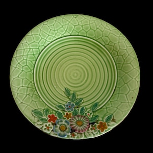 May include: A green ceramic plate with a textured, cracked pattern around the rim and concentric circles in the centre. The plate features a floral design with colourful flowers and green leaves. The plate is set against a black background.