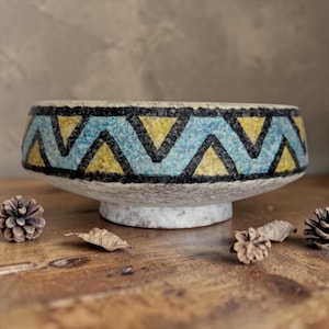 May include: A ceramic bowl with a geometric pattern. The bowl has a wide, shallow shape with a rim decorated with blue, yellow, and black triangles. It rests on a short, textured base. The background is a neutral color.